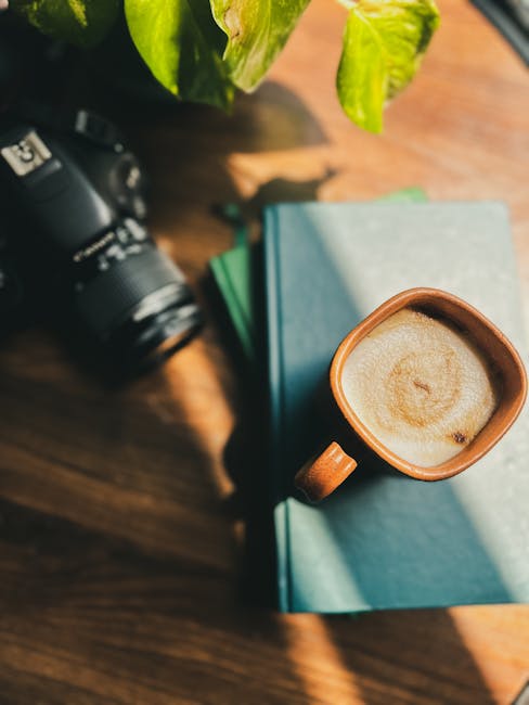 Close-up image of a wooden table surface with a camera partially visible on the left side. In the center, there are two stacked hardcover books with light blue covers, positioned next to a small, brown ceramic mug filled with a frothy coffee or latte, showing light-colored foam on the surface. The scene is illuminated by natural light, casting shadows on the table. A green leafy plant is partially visible at the top left corner, adding a touch of greenery to the setting. This arrangement reflects a casual environment suitable for planning or documenting a house move, aligning with the context of home relocation or furniture transport. The presence of books, a camera, and a beverage suggests a relaxed moment during packing or preparation for a move, commonly handled by services like Man and Van Bounds Green, which specialises in removals and moving logistics.