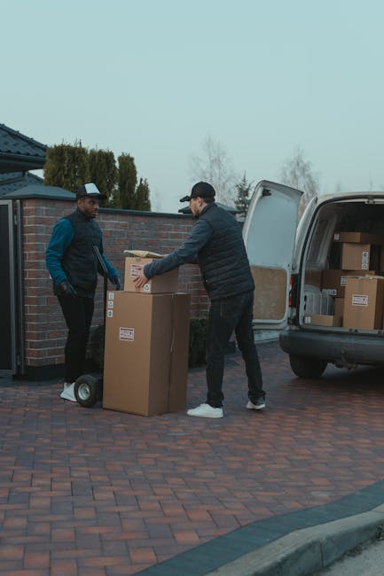 Two men engaged in a home relocation process outside a property on a paved driveway. The man on the left, wearing a black cap, black jacket, and gloves, stands beside a small dolly cart holding a cardboard box. The man on the right, wearing a black cap, dark jacket, and white shoes, is lifting or placing a cardboard box onto a larger box positioned on the ground. Nearby, an open van with its rear doors wide open reveals stacked cardboard boxes inside, ready for transport. The background features a brick wall, a blue-roofed building, and some trees and shrubs under an overcast sky. The scene captures the loading process integral to packing and moving services provided by Man and Van Bounds Green, emphasizing careful handling of furniture and packing materials during house removals.