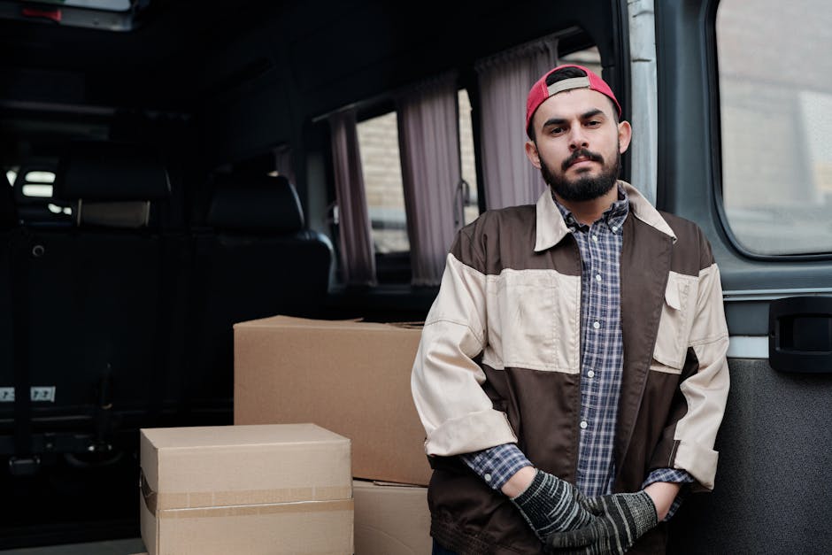 A man with a dark beard, wearing a red cap, beige and brown work jacket over a checked shirt, and grey gloves, stands inside the back of a van during a home relocation process. Behind him are cardboard boxes of various sizes, some sealed with packing tape, placed on the vehicle's floor. The van's interior features black seating and light-colored curtains covering the side windows. The scene takes place outdoors, with a partly visible brick wall and window behind the vehicle. The man appears to be preparing for or engaged in loading or unloading furniture and packing materials as part of a professional removals service offered by Man and Van Bounds Green, specializing in house removals and furniture transport, with a focus on efficient packing and moving logistics.
