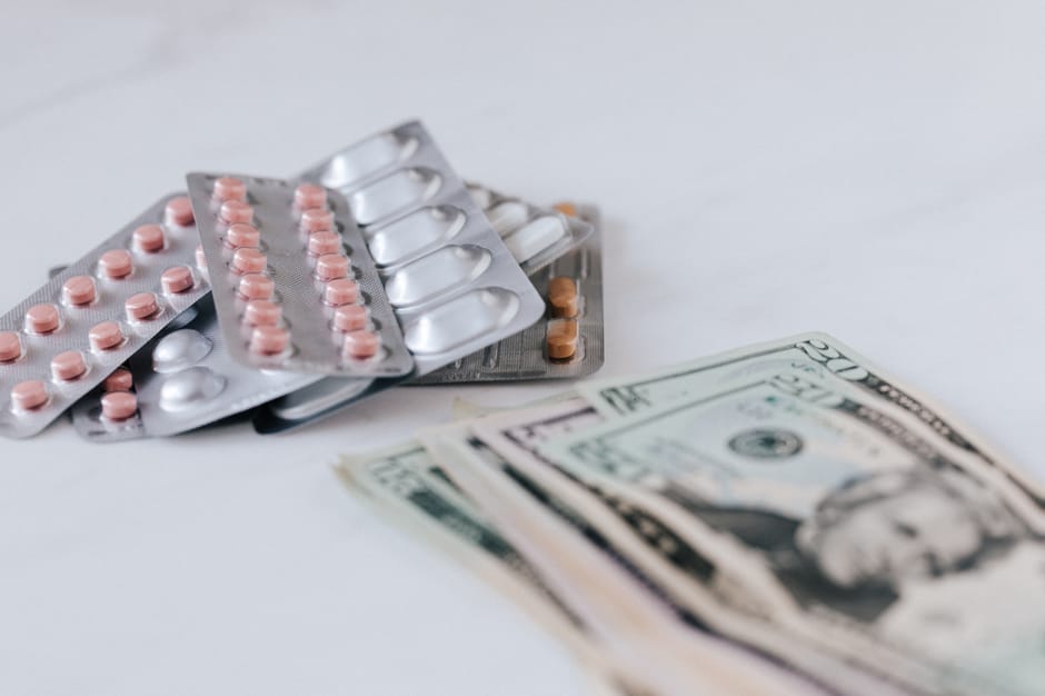 A collection of blister packs containing pink and white pills stacked on top of each other with some tablets still in their foil packaging, placed on a white surface, alongside several US twenty-dollar bills partially visible in the foreground. The blister packs are positioned near the center, with some overlapping, and the money is spread out slightly beneath and adjacent to the medication. The image is captured in natural lighting, emphasizing the textures of the pills and the paper currency. This scene reflects aspects of packing or handling medication during a home relocation process, relevant to furniture transport and moving logistics offered by Man and Van Bounds Green, as seen on their website page about removals costs in Bounds Green.