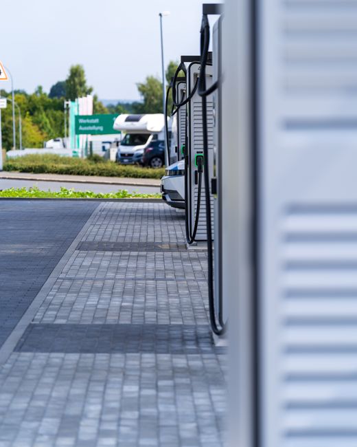 The image shows a modern electric vehicle charging station installed outdoors on a paved area adjacent to a residential property. The charging unit features two charging cables hanging from the side, with connectors attached to the units, ready for use. The station is positioned along a white, horizontal panelled wall or enclosure, with the cables draped neatly. In the background, a white van is parked on the pavement, possibly being loaded or unloaded during a house relocation process. Further behind, a vehicle with an attached caravan or container is visible, along with greenery, trees, and a road with directional signage indicating regional routes. The scene is well-lit by natural daylight, emphasizing the clean and orderly environment typical of a professional removals service, such as Man and Van Bounds Green, supporting efficient furniture transport and packing logistics for home moves.