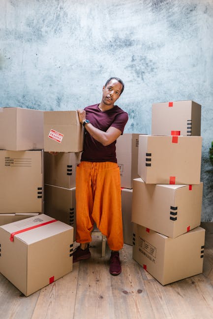 A person wearing an orange pair of trousers and a maroon t-shirt stands indoors, surrounded by multiple cardboard boxes of various sizes, some sealed with red and black packing tape. The individual appears to be in the process of a home or office relocation, with the boxes stacked on a wooden floor against a textured, light blue wall. The scene suggests an organized packing and moving process, likely part of the home relocation services offered by Man and Van Bounds Green. Equipment such as a trolley or lifting straps is not visible, but the individual’s stance indicates handling or preparing boxes for transport. The environment is well-lit, emphasizing the materials used in packing and the spatial arrangement typical of a moving or relocation scenario. This visual context aligns with the customer-focused house removals and furniture transport services provided by [COMPANY_NAME], supporting safe and efficient moving logistics in Bounds Green.
