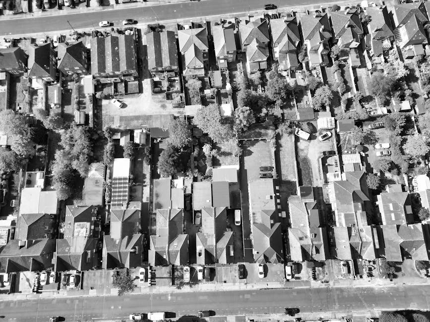 An aerial view of a residential street during winter, showing multiple terraced houses with snow-covered roofs and small backyards enclosed by wooden fences. Cars are parked along both sides of the na