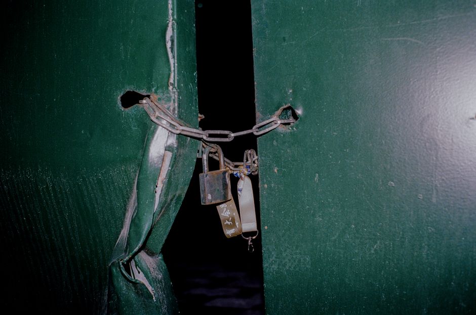 Close-up photograph of a green metal gate secured with a metal chain and a padlock. The chain is looped through a hole in the gate's surface, with the padlock fastened to the chain, ensuring the gate 