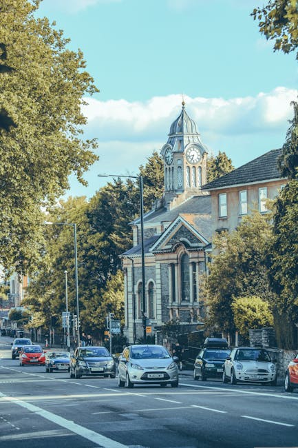 A street scene in Bounds Green during daylight hours, featuring a historical church building with a prominent clock tower and domed spire, surrounded by tall leafy trees and residential buildings. Several cars are parked along the curb and are moving on the road, which has clearly marked lanes and pedestrian crossings. Near the church, a crew from Man and Van Bounds Green is involved in furniture transport or packing activities, with visible moving equipment such as trolleys and blankets. Wooden crates, cardboard boxes, and wrapped furniture pieces are positioned on the pavement and inside the open doorway of a nearby property, indicating a home relocation process. The scene showcases a typical urban environment engaged in essential logistics for house removals, with careful handling of household items and transportation vehicles ready for loading or unloading in the context of urgent or same-day moving services. Bright daylight and natural light enhance visibility, emphasizing the details of the moving operation within the busy neighbourhood.