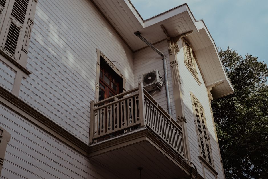 A close-up view of the upper floor of a two-storey house with white wooden siding, featuring a small balcony with a decorative wooden railing. An air conditioning unit is mounted on the exterior wall near a window with brown framing and closed shutters. The house is situated outdoors, with sunlight casting shadows on the facade and a large leafy tree visible on the right side. The photograph captures the building's exterior during daylight, illustrating potential challenges of upper floor access during home relocation or furniture transport processes. This image relates to the services provided by Man and Van Bounds Green, highlighting logistical considerations for removals and moving solutions requiring the lifting and carrying of furniture or appliances to the upper level, especially in properties with limited ground-floor access or staircase constraints.