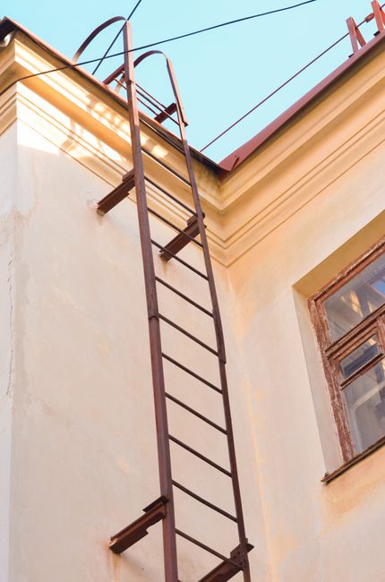 A tall, rusty metal ladder secured to the exterior wall of a two-storey building, extending from the ground level to the roof accessed by a small platform. The building's outer wall is painted in a light cream colour, with a brown window visible on the right side, featuring wooden frames and a partially open windowpane. The ladder is positioned near a window and appears to provide upper floor access, which can pose challenges during house removals or furniture transport when equipment must be carried down or up. The image also shows the edge of a gutter and a section of the roofline under a clear sky, with some wires crossing the top of the frame. Man and Van Bounds Green may use such access points during local moving or packing and moving processes, especially when internal staircases are narrow or unsuitable for furniture passage.