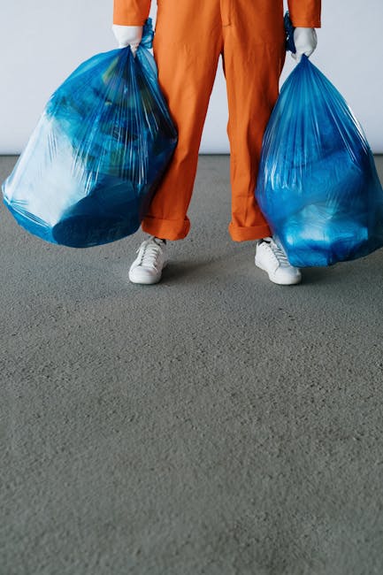 A person dressed in orange work overalls and white sneakers is standing on a grey carpeted floor, holding two large blue plastic garbage bags filled with waste. One bag is held in each hand, with the individual’s legs slightly apart. The background features a plain, light-colored wall, and the lighting is even and neutral. The scene appears to be indoors, possibly in a residential property, during a home relocation or cleaning process. This image relates to waste disposal procedures associated with house removals and end of tenancy cleaning, as managed by companies like Man and Van Bounds Green, assisting with furniture transport and packing during moves.