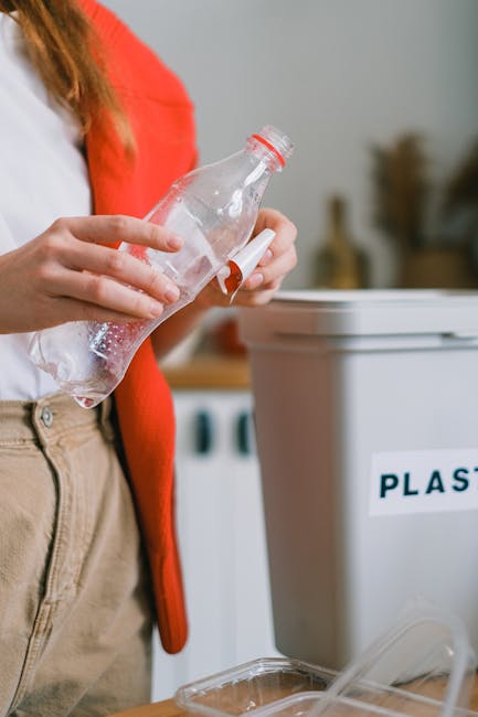 A person wearing a white shirt and beige trousers, with a red cardigan draped over their shoulder, is holding a clear plastic water bottle with a red cap and white label, preparing to dispose of it. They are standing next to a grey plastic recycling bin with a partially visible label reading 'PLAST,' indicating it is designated for plastic waste. The background shows a kitchen setting with a light-colored countertop and blurred objects, suggesting sorting and disposal of recyclable materials as part of home organisation or moving processes. The image captures the act of waste separation, supporting eco-friendly disposal relevant to house removals and packing activities, as managed by Man and Van Bounds Green.