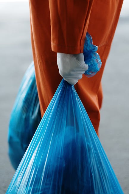 A person dressed in orange work overalls and white sneakers is standing on a grey carpeted floor, holding two large blue plastic garbage bags filled with waste. One bag is held in each hand, with the individual’s legs slightly apart. The background features a plain, light-colored wall, and the lighting is even and neutral. The scene appears to be indoors, possibly in a residential property, during a home relocation or cleaning process. This image relates to waste disposal procedures associated with house removals and end of tenancy cleaning, as managed by companies like Man and Van Bounds Green, assisting with furniture transport and packing during moves.