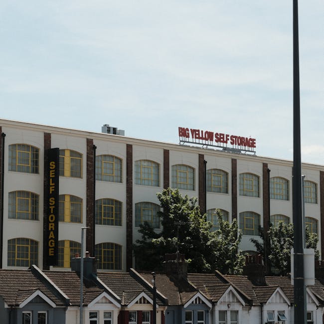 A multi-story industrial-style building with a white facade and large yellow-framed arched windows, featuring vertical black and yellow signage indicating 'Self Storage.' On the roof, a red illuminated sign reads 'Big Yellow Self Storage.' In front of the building, there are several small residential houses with pitched roofs and chimneys, partially obscured by green leafy trees. The sky above is partly cloudy with patches of blue, and a tall street lamp is visible on the right side of the image. The scene suggests a commercial storage facility located in an urban area, potentially used for home relocation or furniture transport services, as indicated by the context of house removals.
